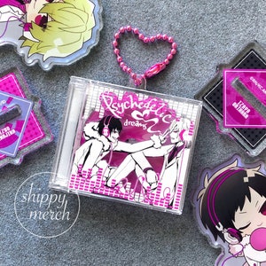May include: A clear plastic CD case with a pink and white graphic featuring two anime-style characters and the text "Psychedelic dreams." A pink heart-shaped keyring is attached. Other anime-style merchandise is also visible.