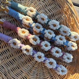 May include: Close-up of several macrame keychains in a woven basket. Each keychain features a different pastel color, including blue, green, purple, and pink. The keychains are adorned with white daisy flower accents and gold-colored clasps.