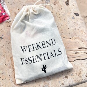 Może przedstawiać: Biała bawełniana torba ze sznurkiem, z nadrukiem "Weekend Essentials" i małym kaktusowym wzorem.