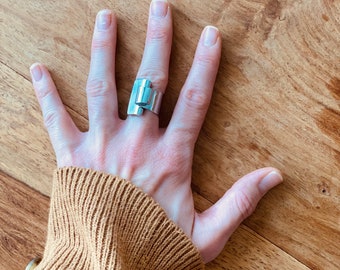 May include: A silver ring with a geometric design. The ring is worn on the middle finger of a hand with a brown sweater sleeve.