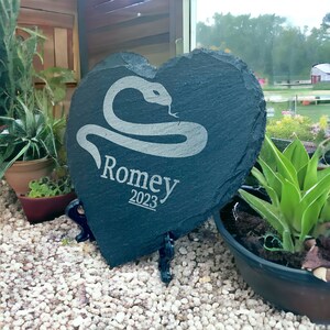 May include: A black heart-shaped slate stone with a white snake design and the name "Romey" and the year "2023" engraved on it. The stone is on a black stand and is sitting on a bed of white gravel.
