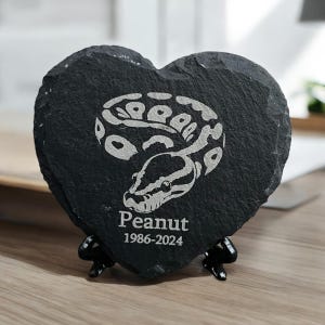May include: A heart-shaped black slate coaster featuring a white snake design and the name "Peanut" with the years 1986-2024. The coaster is supported by a black metal stand. The coaster is made of dark grey slate.