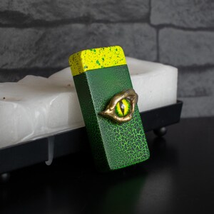 Op de afbeelding: Een groen en geel rechthoekig object met een textuuroppervlak en een goudkleurige, oogvormige versiering. Het object rust op een wit blok en een zwart dienblad, tegen een grijze bakstenen muur.