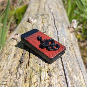 May include: A black and red lighter with a snake design on the front. The lighter is sitting on a wooden log.