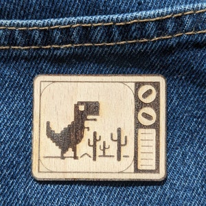 Könnte beinhalten: Ein Holzausschnitt des klassischen Google Chrome Dinosaurierspiels. Der Dinosaurier rennt durch eine Wüstenlandschaft mit Kakteen. Der Ausschnitt befindet sich in einem Retro-Fernsehrahmen mit einer Pause-Taste und einer Lautstärkeregelung.