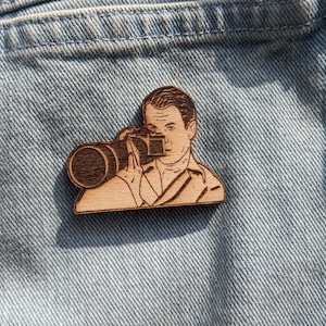 Puede incluir: Un pin de madera con una ilustración en blanco y negro de un hombre sosteniendo una cámara. El hombre lleva una camisa de cuello y tiene el pelo corto. El pin está sobre un fondo de mezclilla azul.