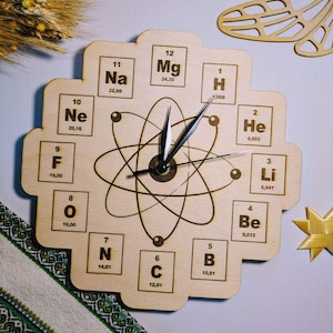 May include: Wooden wall clock featuring the periodic table of elements, with an atom design in the center. The clock face includes element symbols, atomic numbers, and atomic weights. The clock hands are black.