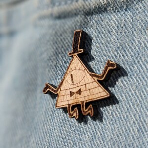 May include: A wooden pin of Bill Cipher, a character from the animated series Gravity Falls. Bill Cipher is a triangular-shaped demon with a single eye, a bow tie, and a top hat. The pin is brown and black.