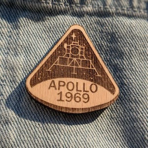 Puede incluir: Un pin de madera en forma de triángulo con una imagen grabada con láser del módulo lunar Apollo 11 aterrizando en la luna. El pin también incluye el texto "APOLLO 1969".