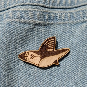 Può includere: Uno spillo di legno a forma di pesce con un design marrone scuro e bianco. Lo spillo è su uno sfondo di denim blu.