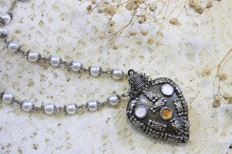 May include: A silver heart-shaped pendant necklace with a pearl chain. The pendant is decorated with small silver flowers and round mirrored accents.