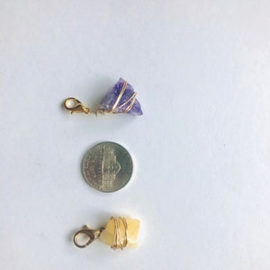 K&ouml;nnte beinhalten: Zwei goldumwickelte Kristall-Anh&auml;nger mit einem goldenen Verschluss. Die Anh&auml;nger sind etwa so gro&szlig; wie eine US-amerikanische 25-Cent-M&uuml;nze. Der gr&ouml;&szlig;ere Anh&auml;nger ist lila und der kleinere ist gelb.