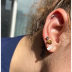 May include: A person wearing three earrings in their left ear. The earrings are made of different materials and shapes. The top earring is a small silver stud. The middle earring is a small gold triangle. The bottom earring is a long, thin, white crystal.