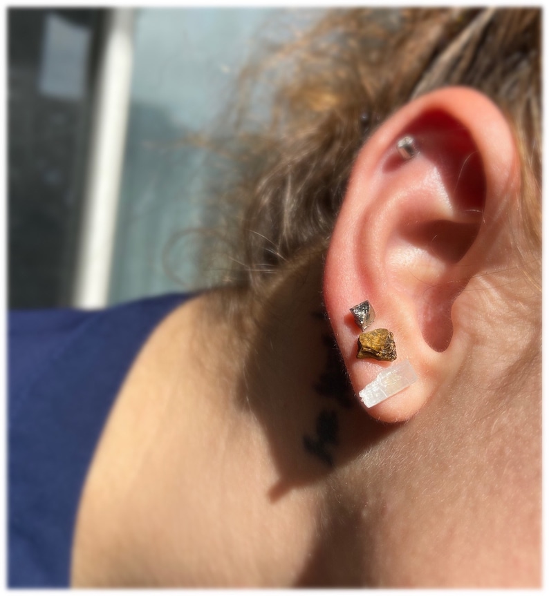 May include: A person wearing three earrings in their left ear. The earrings are made of different materials and shapes. The top earring is a small silver stud. The middle earring is a small gold triangle. The bottom earring is a long, thin, white crystal.
