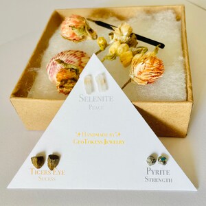 May include: Three pairs of earrings in a triangular box. The earrings are made of selenite, tiger's eye, and pyrite. The box is white with black text that reads "Handmade by GeoTokens Jewelry". The selenite earrings are labeled "Peace", the tiger's eye earrings are labeled "Success", and the pyrite earrings are labeled "Strength".
