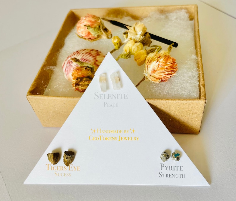 May include: Three pairs of earrings in a triangular box. The earrings are made of selenite, tiger's eye, and pyrite. The box is white with black text that reads "Handmade by GeoTokens Jewelry". The selenite earrings are labeled "Peace", the tiger's eye earrings are labeled "Success", and the pyrite earrings are labeled "Strength".