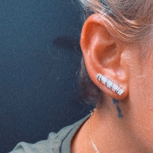May include: A close-up of a person's ear with a silver ear cuff featuring multiple small white stones. The ear cuff is worn on the upper part of the ear.