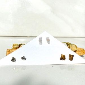 May include: Three pairs of earrings displayed on a white triangular card. The earrings are made of different materials, including clear crystal, silver, and wood.