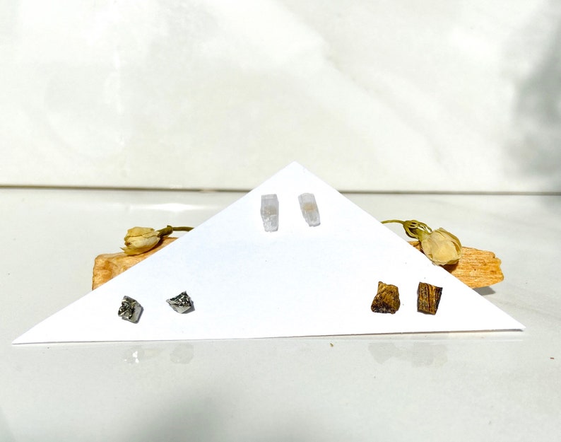 May include: Three pairs of earrings displayed on a white triangular card. The earrings are made of different materials, including clear crystal, silver, and wood.