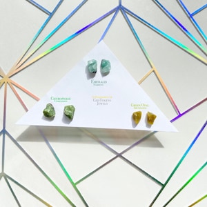 May include: Four pairs of raw, uncut gemstone earrings. The earrings are labeled with their names and properties: Emerald, Harmony; Green Opal, Abundance; Chrysoprase, Compassion; and Green Opal, Abundance. The earrings are displayed on a white card with the text "Handmade at GeoTokens Jewelry".