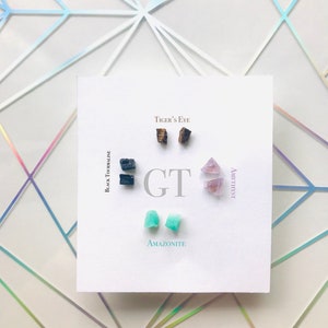 May include: A set of six gemstone earrings, including black tourmaline, tiger's eye, amethyst, and amazonite. The earrings are displayed on a white card with the names of the gemstones written below each pair.