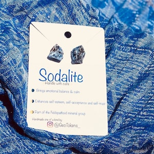 May include: A pair of blue sodalite crystal earrings on a white card with the text "Sodalite Handle with care" and a description of the stone's properties. The card also includes the text "Handmade one of a kind by: @GeoTokens_"