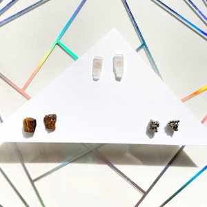 May include: Three pairs of earrings featuring different natural stones. The first pair is made of clear quartz crystals, the second pair is made of brown tiger's eye stones, and the third pair is made of silver pyrite stones.