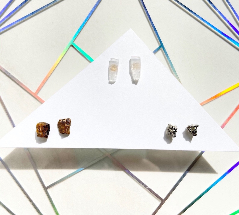 May include: Three pairs of earrings featuring different natural stones. The first pair is made of clear quartz crystals, the second pair is made of brown tiger's eye stones, and the third pair is made of silver pyrite stones.