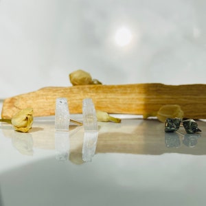 May include: A pair of selenite crystal earrings with gold posts. The crystals are clear and have a striated texture. The earrings are displayed on a wooden surface with dried flowers.