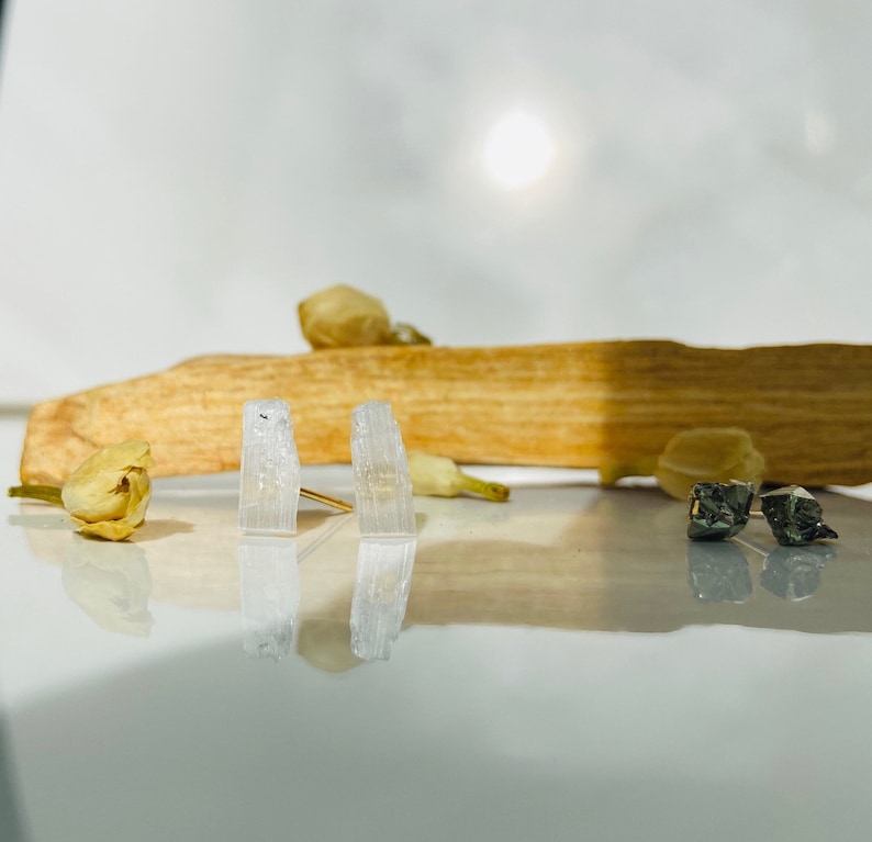 May include: A pair of selenite crystal earrings with gold posts. The crystals are clear and have a striated texture. The earrings are displayed on a wooden surface with dried flowers.