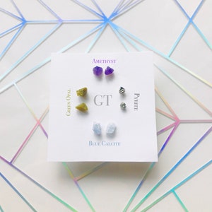 May include: A set of six gemstone earrings, including amethyst, green opal, pyrite, and blue calcite. The earrings are displayed on a white card with the names of the stones written below each pair.