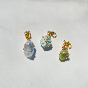K&ouml;nnte beinhalten: Drei kleine, nat&uuml;rliche Stein-Anh&auml;nger, die mit goldfarbenem Draht umwickelt sind. Die Steine sind hellblau, t&uuml;rkis und gr&uuml;n. Die Anh&auml;nger sind an goldfarbenen Karabinerhaken befestigt. Der Text "Calming Set" steht unter den Anh&auml;ngern.