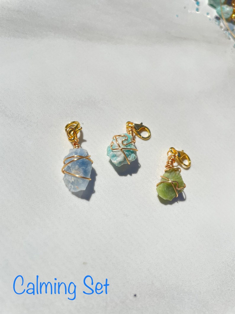 K&ouml;nnte beinhalten: Drei kleine, nat&uuml;rliche Stein-Anh&auml;nger, die mit goldfarbenem Draht umwickelt sind. Die Steine sind hellblau, t&uuml;rkis und gr&uuml;n. Die Anh&auml;nger sind an goldfarbenen Karabinerhaken befestigt. Der Text "Calming Set" steht unter den Anh&auml;ngern.