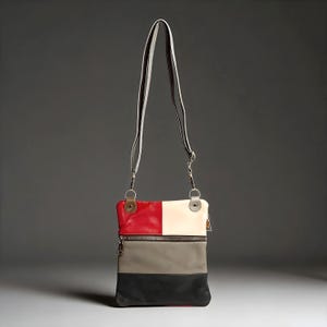 May include: A rectangular crossbody bag featuring a color-blocked design with red and cream panels on the top, a gray middle section, and a black base. The bag includes a long, adjustable shoulder strap attached with silver-toned metal hardware.