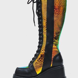 May include: A knee-high lace-up boot featuring a black matte finish combined with iridescent, gold-toned snakeskin-textured panels. The boot includes a thick, black platform wedge sole with a deep-tread lug design and black laces.
