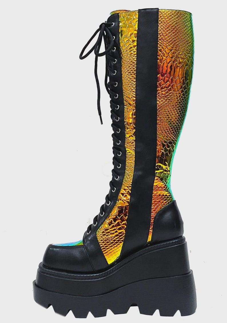 May include: A knee-high lace-up boot featuring a black matte finish combined with iridescent, gold-toned snakeskin-textured panels. The boot includes a thick, black platform wedge sole with a deep-tread lug design and black laces.
