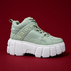 May include: A sage green high-top sneaker featuring a textured leather upper, matching laces, and a thick, chunky white rubber platform sole with deep grooves. The side panel includes rectangular accents and a small metallic silver detail.