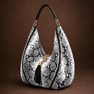 May include: A hobo-style handbag featuring a black and white snakeskin pattern. The bag includes a black top handle, gold-tone hardware, and a decorative gold chain accent with a black tassel hanging from the center of the front panel.