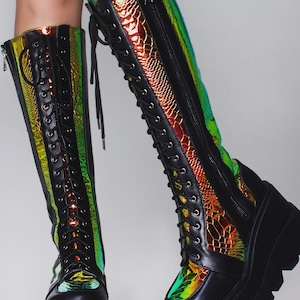 May include: Knee-high boots featuring a black matte finish with iridescent, holographic snake-patterned panels. These boots include a front lace-up closure with black eyelets, a side zipper, and a thick, chunky black platform lug sole.