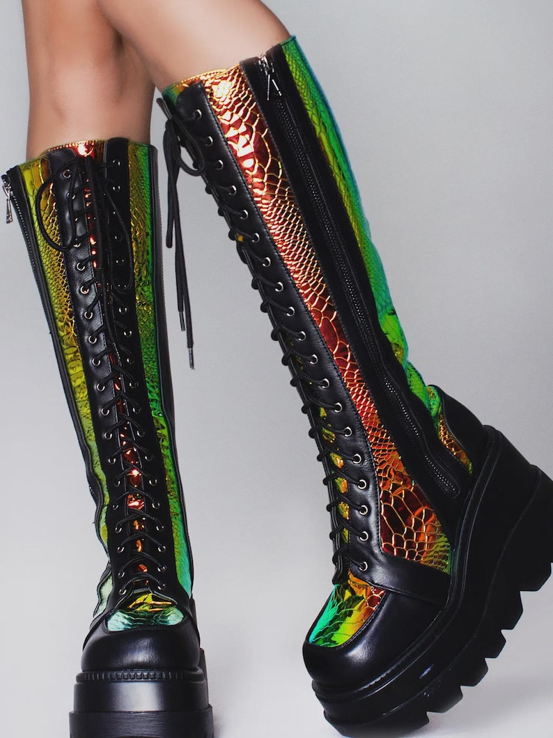 May include: Knee-high boots featuring a black matte finish with iridescent, holographic snake-patterned panels. These boots include a front lace-up closure with black eyelets, a side zipper, and a thick, chunky black platform lug sole.