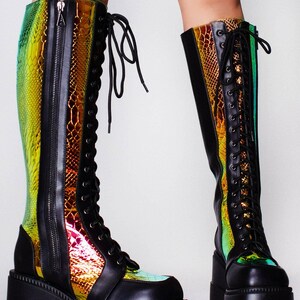 May include: Knee-high platform boots featuring a black matte finish with iridescent, snake-patterned panels that shift between green, gold, and red. These boots include a front lace-up closure with black eyelets and a full-length side zipper.