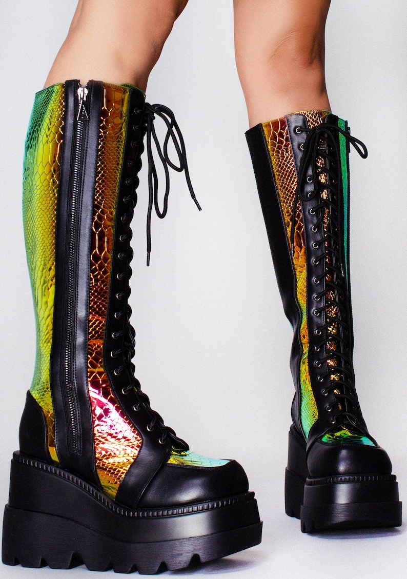 May include: Knee-high platform boots featuring a black matte finish with iridescent, snake-patterned panels that shift between green, gold, and red. These boots include a front lace-up closure with black eyelets and a full-length side zipper.