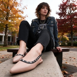 May include: A pair of beige pointed-toe slingback flats featuring a black trim adorned with silver-toned pyramid studs. The shoes have a thin adjustable ankle strap with a small metal buckle and a low, flat heel, showcasing a sleek and structured design.