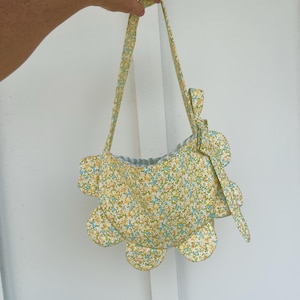 May include: A floral print handbag with a distinctive scalloped edge. The bag displays a yellow, blue, and white floral pattern. It has long straps and is held by a hand.