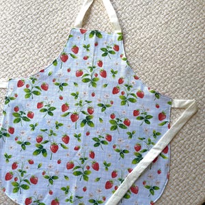May include: A light blue apron with a repeating pattern of red strawberries, green leaves, and white flowers. The apron has white ties and a neck strap, perfect for kitchen use.