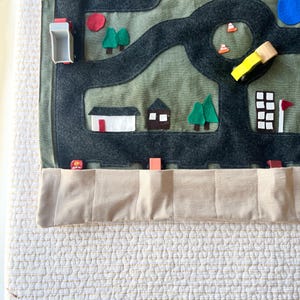 Puede incluir: Un tapete de juego de fieltro con un mapa de carreteras con un diseño de carretera gris oscuro sobre un fondo verde. El tapete presenta recortes de fieltro de edificios, árboles y un símbolo 'H' azul. Un bolsillo beige está cosido en la parte inferior.