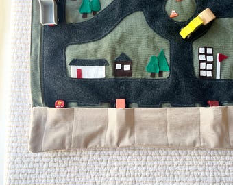 May include: A felt road map play mat with a dark gray road design on a green background. The mat features felt cutouts of buildings, trees, and a blue 'H' symbol. A beige pocket is sewn at the bottom.