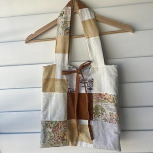 May include: A patchwork tote bag with a brown leather tie closure. The bag is made of various fabric squares in shades of white, tan, and brown, some with floral and landscape patterns. The bag hangs from a wooden hanger.