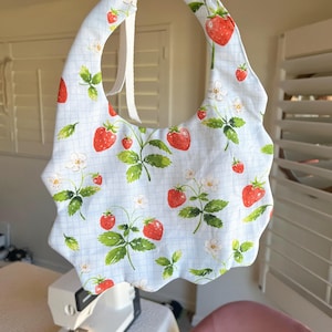 May include: A baby bib with a light blue background and a pattern of red strawberries, green leaves, and white flowers. The bib has a scalloped edge and white ties. The bib is held up by a hand.