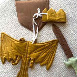 May include: A mustard yellow dragon wing set, a brown shield, a mustard yellow crown, and a brown sword with a green handle are arranged on a white quilted surface. The dragon wings are the focal point, with detailed stitching.